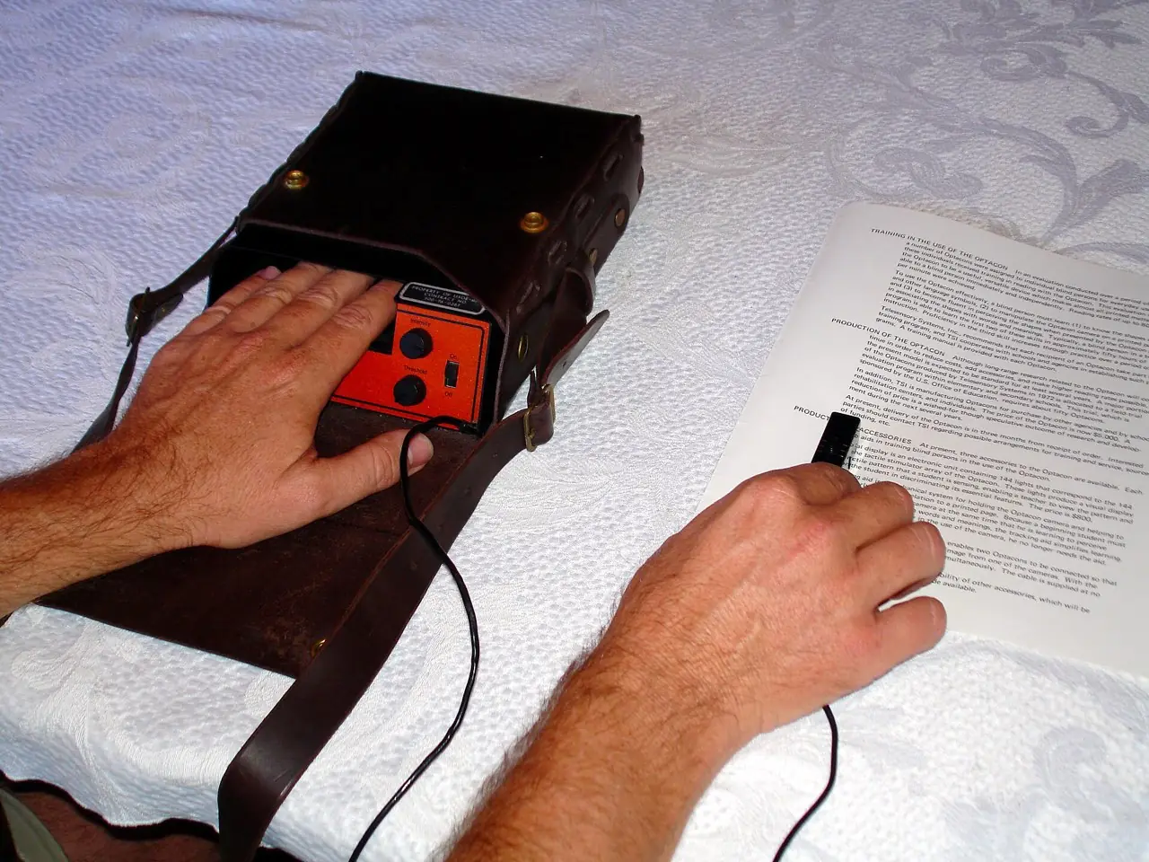 Two hands using an Optacon: one hand rests on the reading pad while the other guides a small camera along a line of printed text.